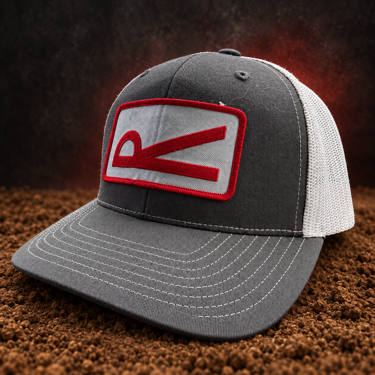 Gray and white Rabon trucker hat with red embroidered patch on an arena-dirt studio backdrop