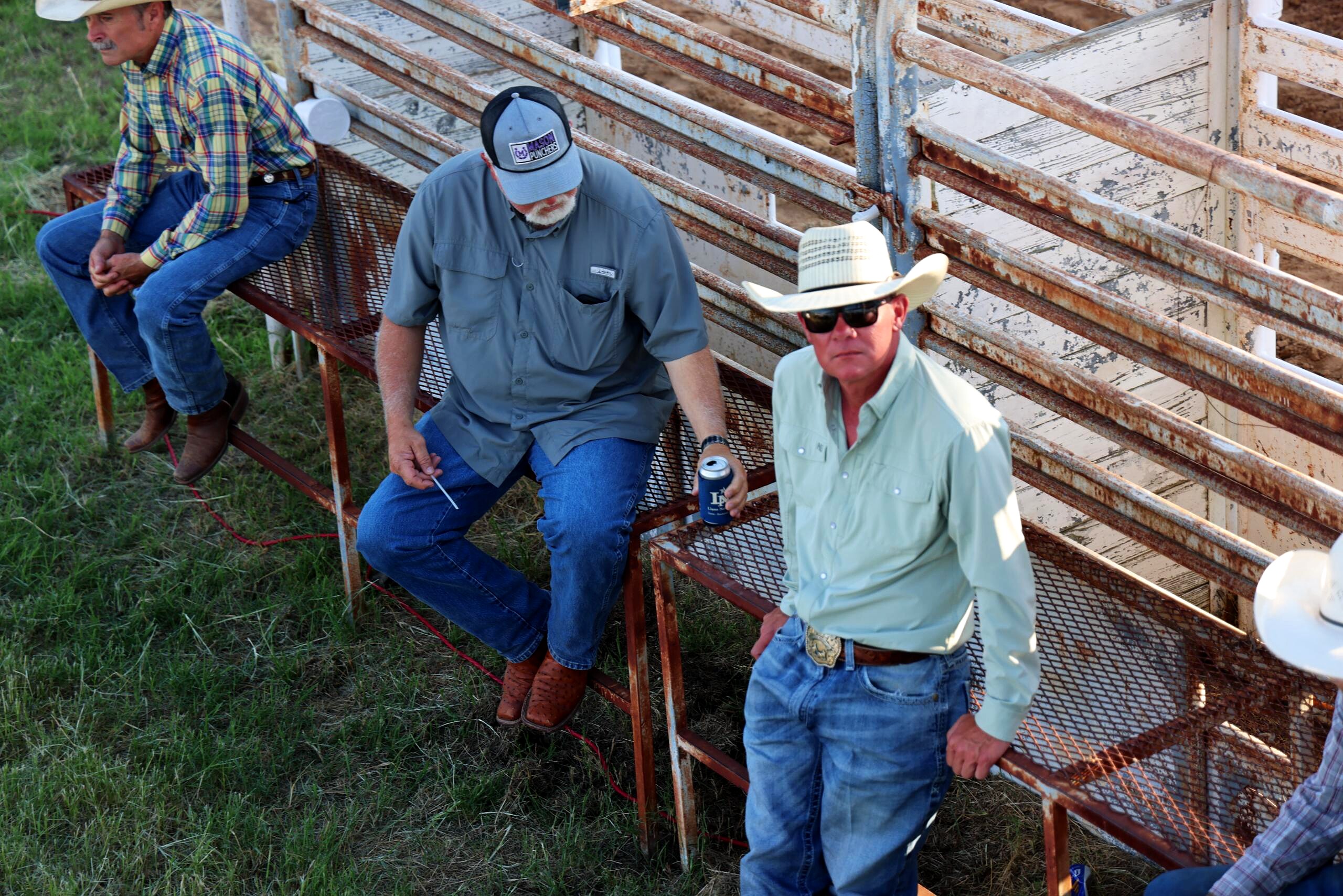 Three men in cowboy hats seated near rusty arena pens